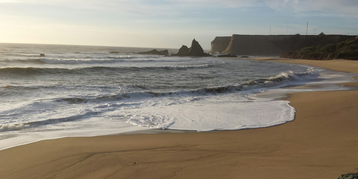 Ocean waves roll in on golden sand with a backdrop of the sharkfin rock at Martin's Beach