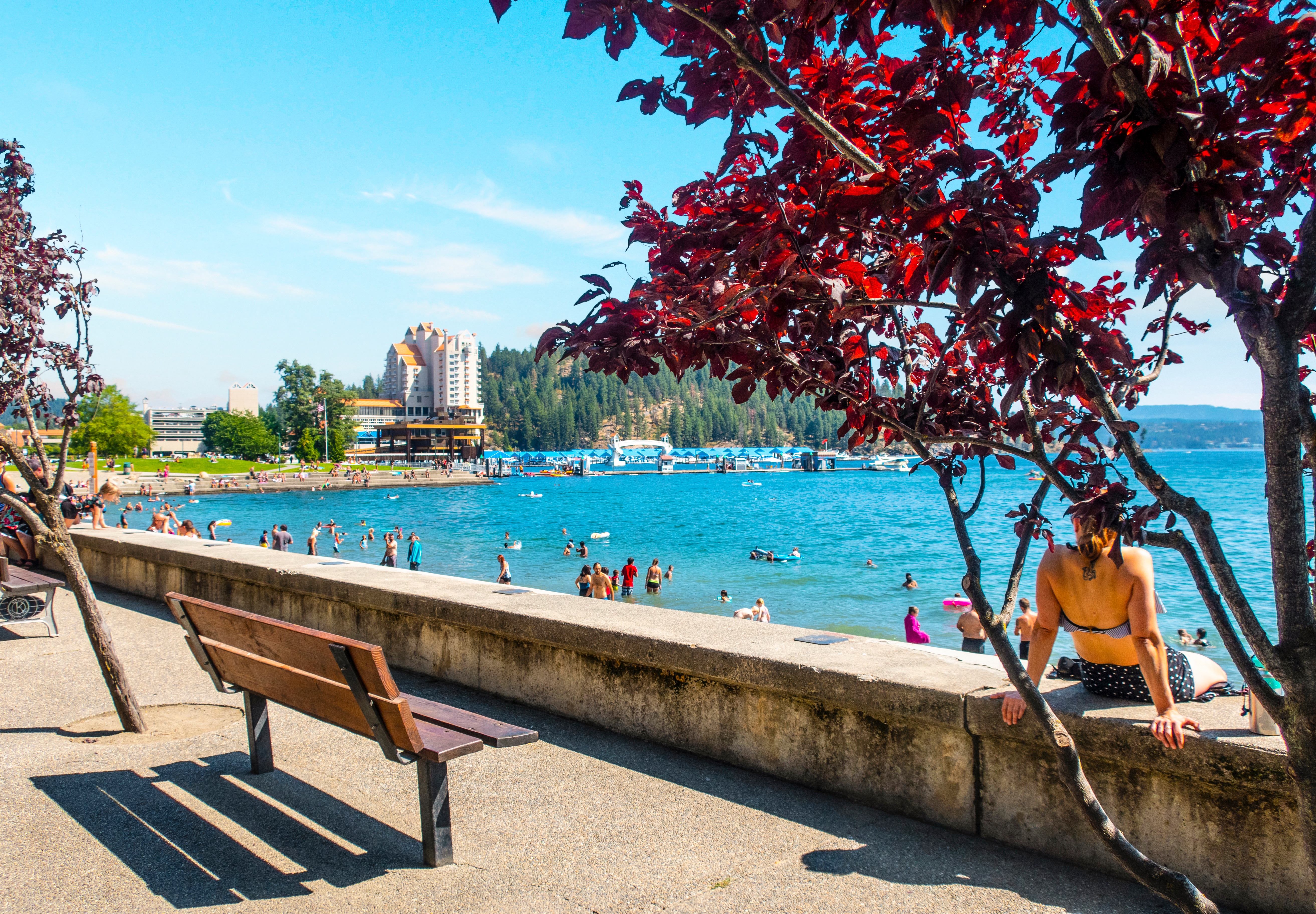 a-woman-relaxes-on-a-barrier-wall-at-the-city-beach-and-park-along-the-shores-of-lake-coeur-dalene-idaho-on-a-busy-summer-day