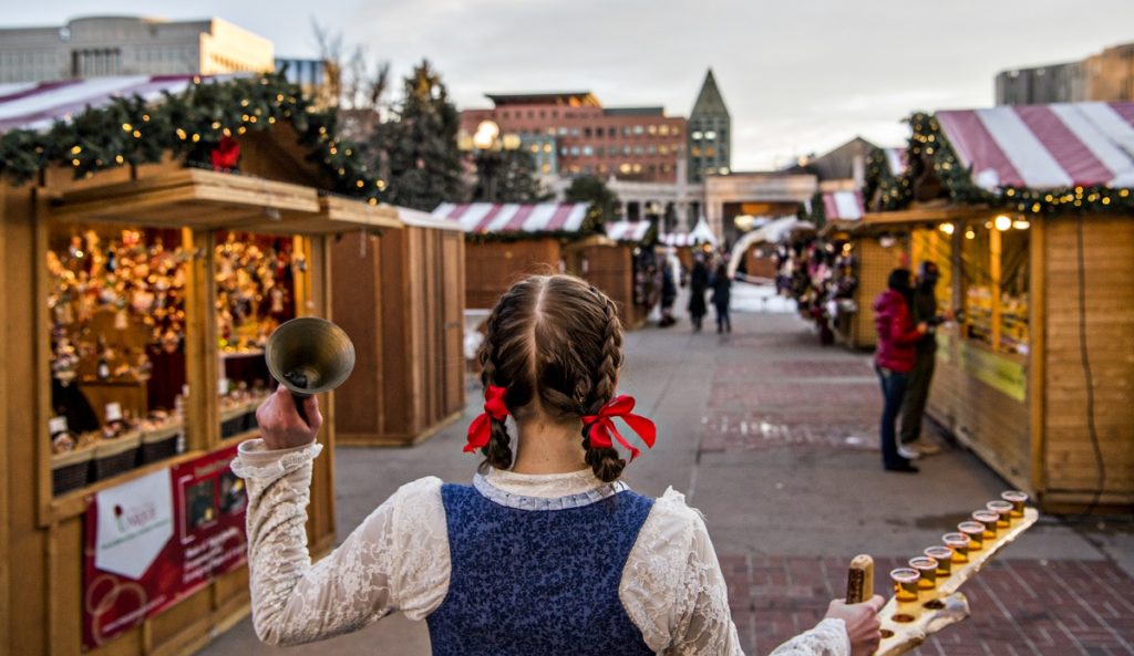 woman with bell and row of shots walks down alley of vendors