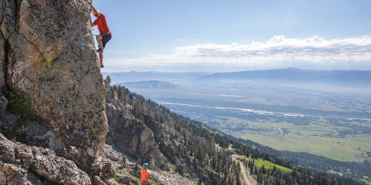 person climbs a rock face with a river and hills in background