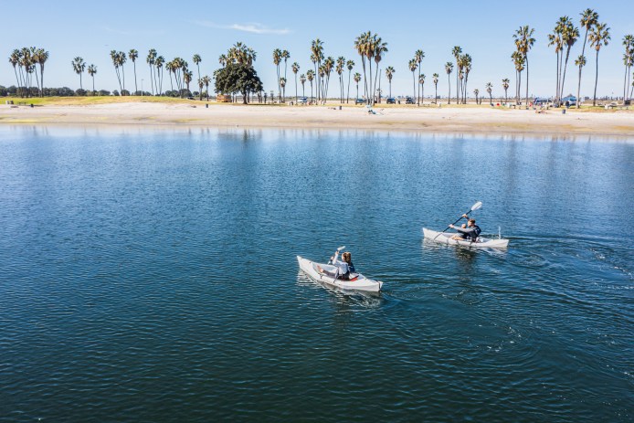 two people in kayaks on smooth water, beach and palm trees in distance