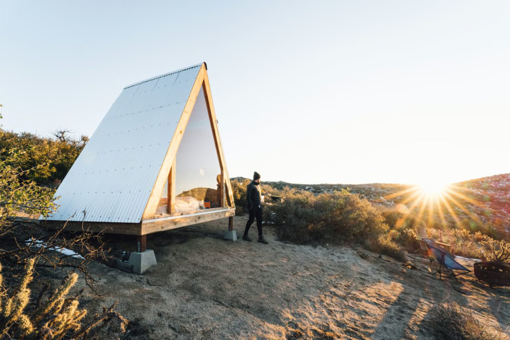 man standing in front of a frame cabin at sunset