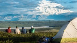 four people sitting near a tent on a hill, looking at a distant, snowy mountain