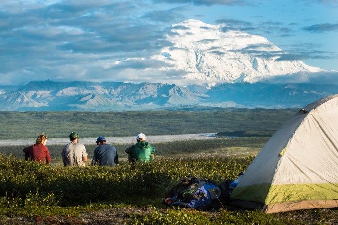 four people sitting near a tent on a hill, looking at a distant, snowy mountain