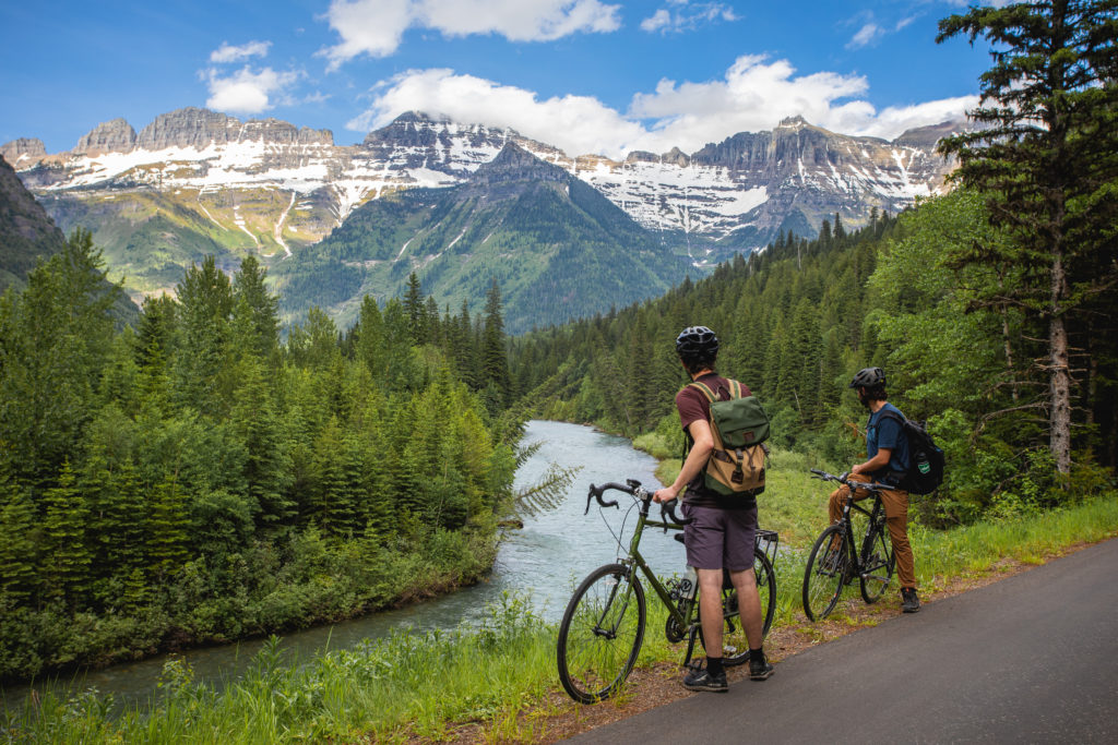 two cyclists stand beside bikes looking out at a river and mountains