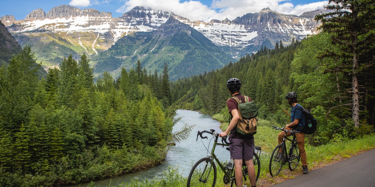 two cyclists stand beside bikes looking out at a river and mountains